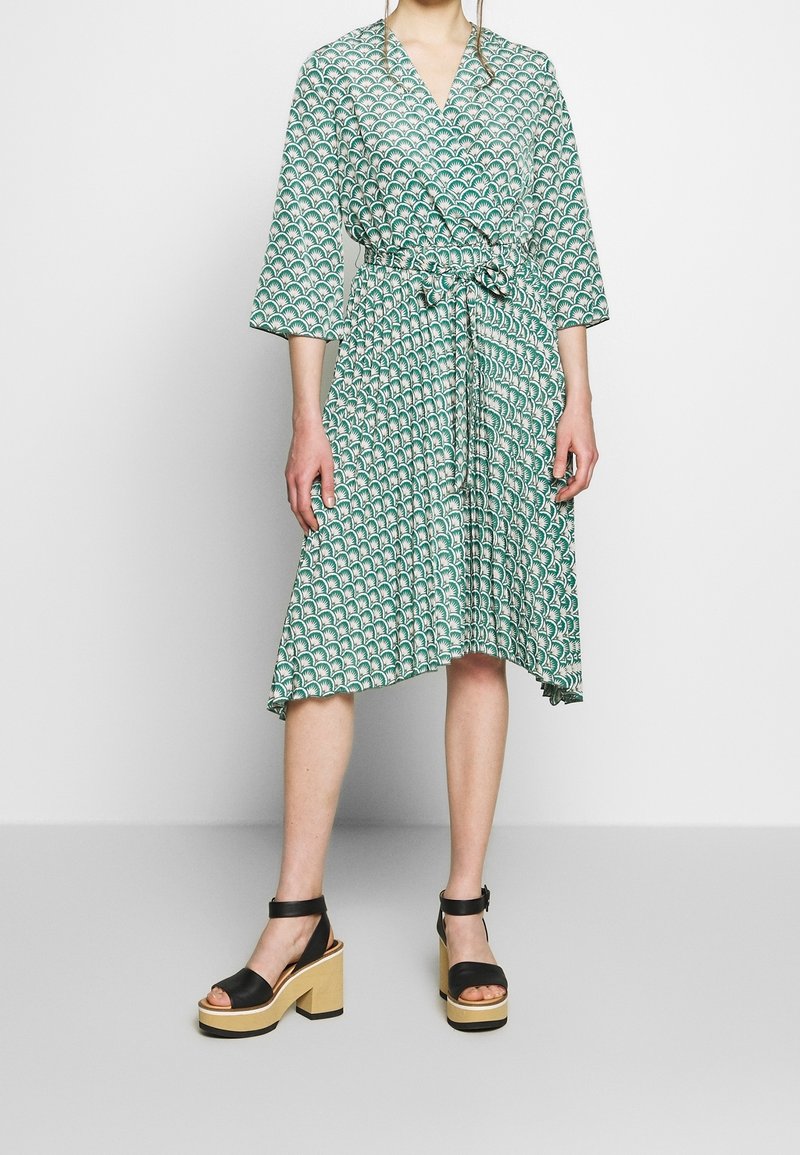 Femme portant une robe portefeuille verte et blanche à motifs avec des manches 3/4 et des sandales à plateforme noires avec des brides chevilles, se tenant devant un fond uni.