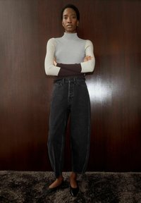 Personne debout, bras croisés, portant un pull à col roulé bicolore, un jean noir taille haute et des ballerines noires pointues devant un mur en bois.