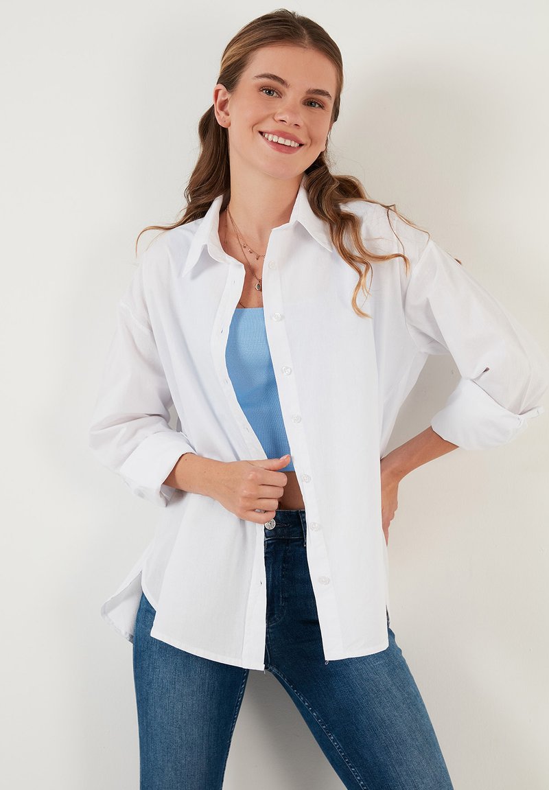 White button-up shirt with long sleeves, worn over a light blue tank top, paired with dark blue jeans. Simple design, fabric appears smooth.
