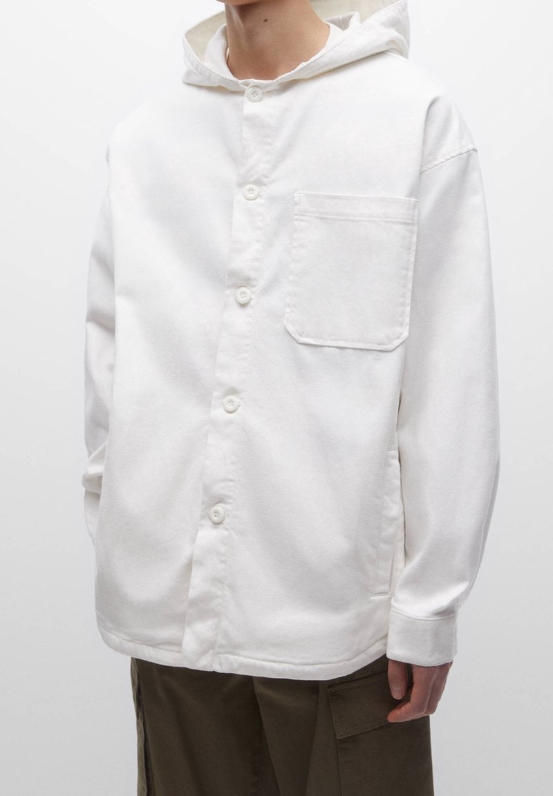 Camisa blanca con capucha, hecha de tela suave, que cuenta con un cierre de botones en la parte delantera y un solo bolsillo en el pecho. Mangas largas con puños.