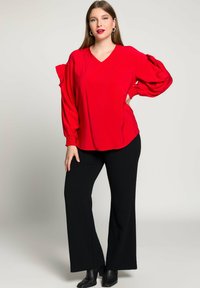 Blusa rossa a V con maniche a sbuffo e polsini aderenti, abbinata a pantaloni neri a gamba larga e stivali alla caviglia, su uno sfondo neutro.