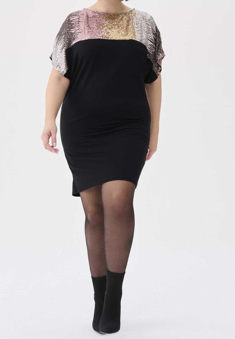 Žena kráčející v černých šatech s lesklou flitrovou horní částí, v průhledných černých punčochách a černých kotníkových botách na bílém pozadí.