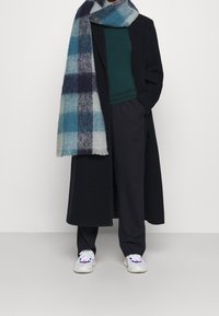 Lang sort frakke, teal top, grå og blå plaid tørklæde, løse sorte bukser og hvide sneakers med farverige accenter.