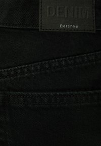 Svart denimtyg med en texturerad yta, utrustad med en rektangulär läderetikett präglad med "DENIM" och "Bershka" i silvertext.