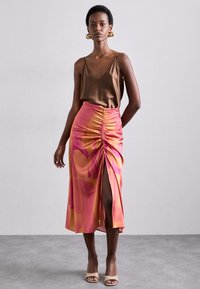 Camisole en soie beige associé à une jupe en satin rose et orange à motifs, avec fronces et une fente sur le côté. Des sandales à talons beiges complètent le look.