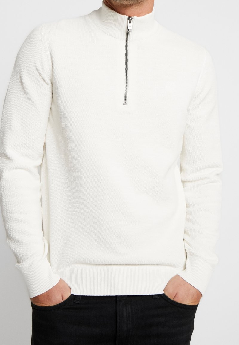 Maglione bianco a maglia con collo alto, dotato di una cerniera argentata, polsini a costine e un motivo testurizzato. Indossato sopra un paio di jeans neri.