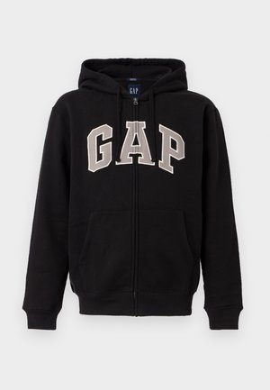 Melns kapučjakas ar rāvējslēdzēju, priekšpusē kabatas un krēmkrāsas "GAP" uzraksts uz krūtīm, ar kapuci un auklu.