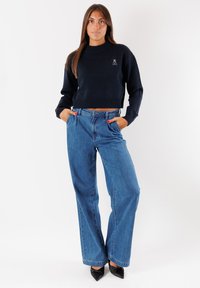 Maglione corto blu navy con collo e orlo a coste, abbinato a jeans blu a vita alta e gamba dritta. Il modello indossa décolleté nere con punta appuntita.