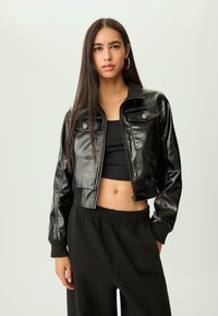 Mlada žena s dugom tamnom kosom odjevena u crnu kožnu jaknu, crni crop top, crne hlače i zlatne okrugle naušnice na jednobojnoj pozadini.
