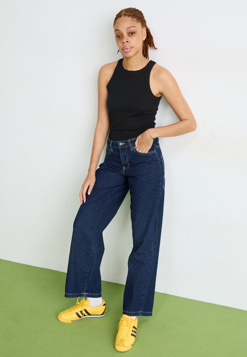 Top nero senza maniche abbinato a jeans in denim blu a vita alta e gamba larga, con sneakers gialle brillanti con strisce nere. Sfondo verde.