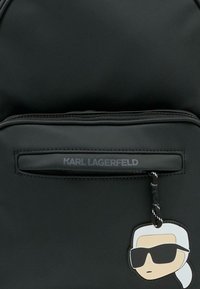 Czarny plecak z gładkiego materiału, z przednią kieszenią na zamek błyskawiczny z logo "KARL LAGERFELD" oraz detalem w formie przywieszki w stylu kreskówkowym.