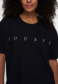 Camiseta de algodón negra de manga corta, con cuello redondo, que presenta el texto "10 DÍAS" en blanco, centrado en el frente. Textura suave, corte relajado.