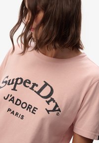 Camiseta de algodón rosa con un estampado gráfico negro que incluye "SuperDry J'ADORE PARIS". Mangas cortas y cuello redondo.