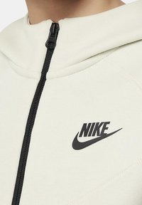Sweat à capuche beige clair zippé en tissu doux, avec une fermeture éclair noire et le logo Nike noir sur la poitrine.