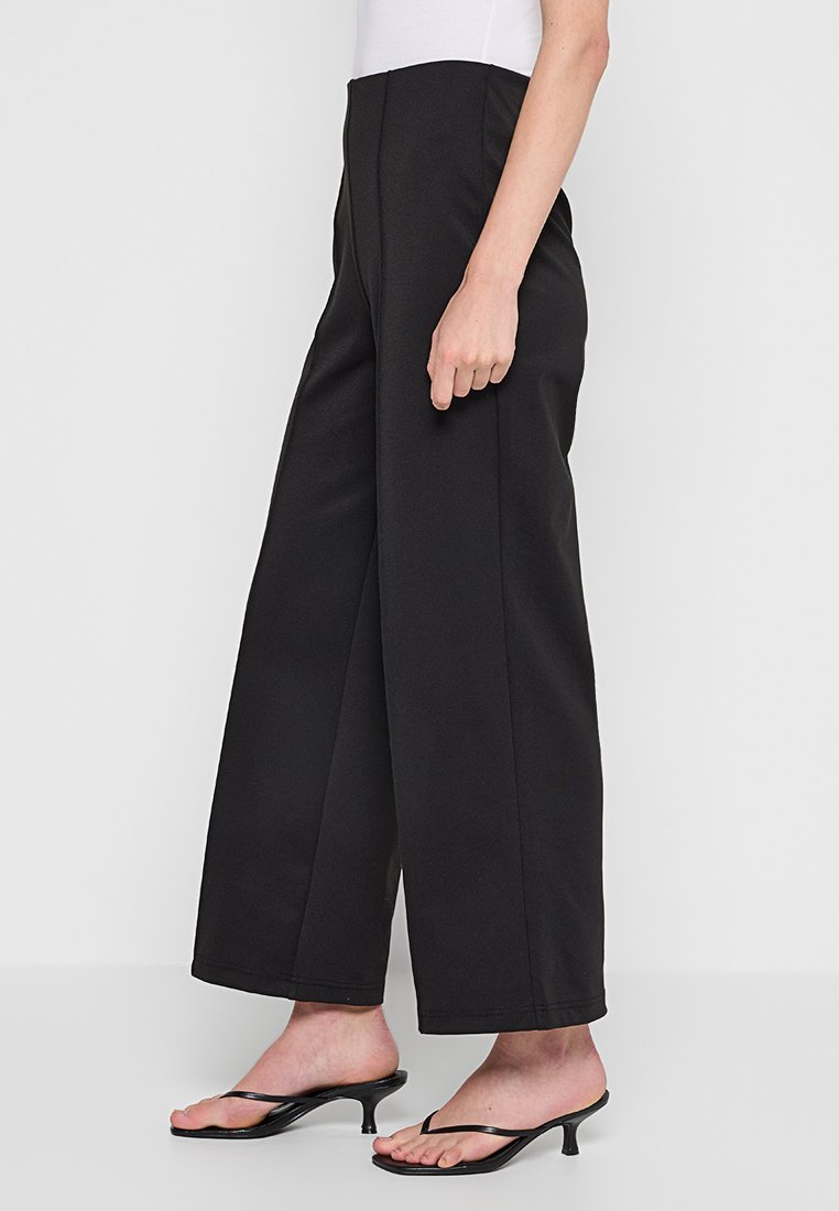 Czarne szerokie spodnie z gładkiego materiału, z wysokim stanem i plisami z przodu. Połączone z czarnymi sandałami na obcasie.