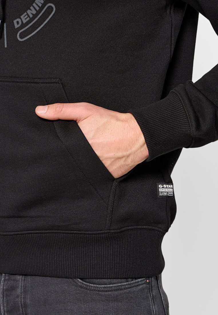 Sudadera negra con bolsillo frontal, puños y dobladillo acanalados, que presenta un estampado "DENIM" en el pecho y una pequeña etiqueta de marca en el lado inferior.