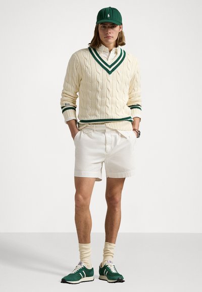 Creme kabelstrikket sweater med grønne accenter, hvide shorts, grønne sneakers og en grøn hat. Rund halsudskæring og struktureret design.