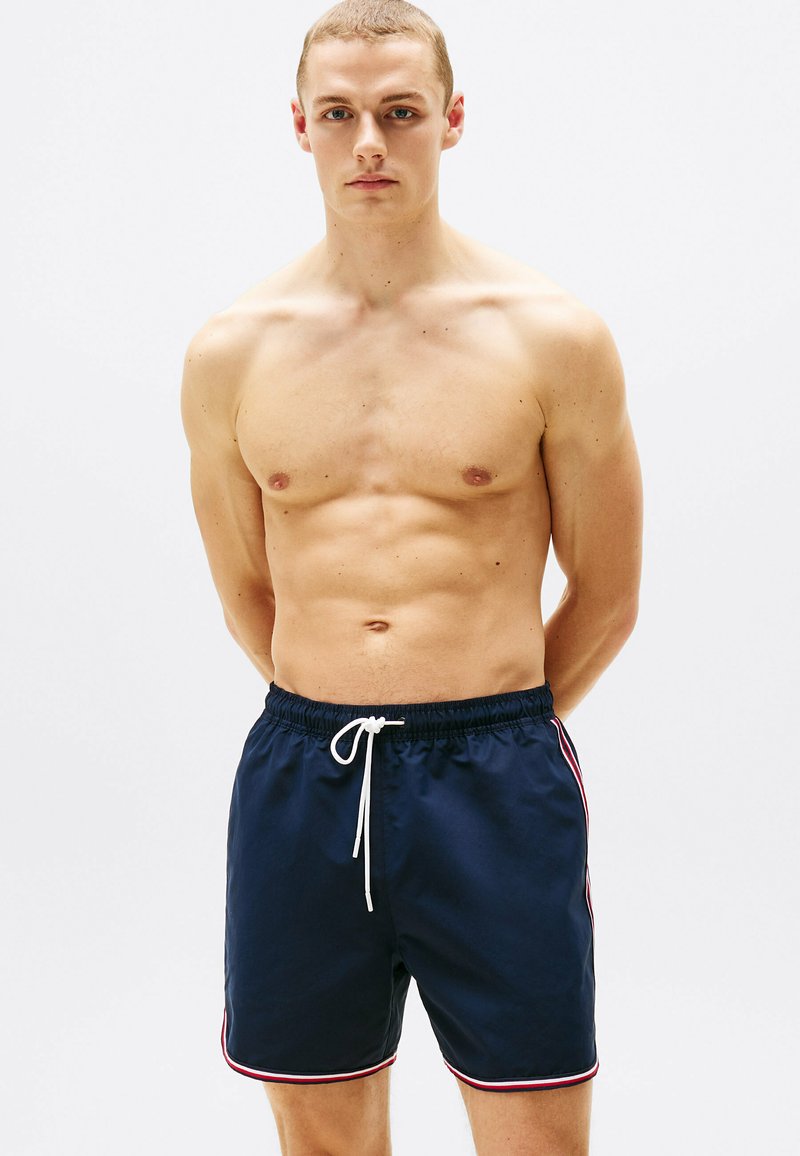 Bañadores de color navy con cintura ajustable, con franjas laterales en rojo y blanco. Fabricados en un tejido suave, de longitud a medio muslo, con un corte casual.