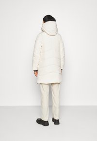 Lungo cappotto imbottito crema con cappuccio, caratterizzato da un design a zigzag trapuntato e una texture morbida, abbinato a pantaloni crema e scarpe nere.