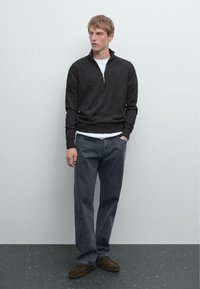 Un pull gris foncé avec un col zippé, superposé sur une chemise blanche. Associé à un pantalon en jean sombre ample et des chaussures en mocassin marron.