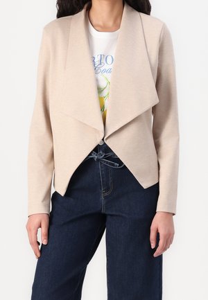 Veste beige drapée ouverte sur un t-shirt blanc graphique, associée à un jean large taille haute bleu foncé avec ceinture nouée.