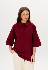 Bordeaux oversized shirt met een puntige kraag, korte mouwen en een zachte katoentextuur. Gepaard met lichtbeige broeken.
