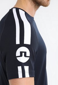 Marineblaues Sportshirt mit weißen Streifen auf den Schultern, strukturiertem Stoff, rundem Logo auf dem Ärmel und tailliertem Schnitt für verbesserte Bewegungsfreiheit.