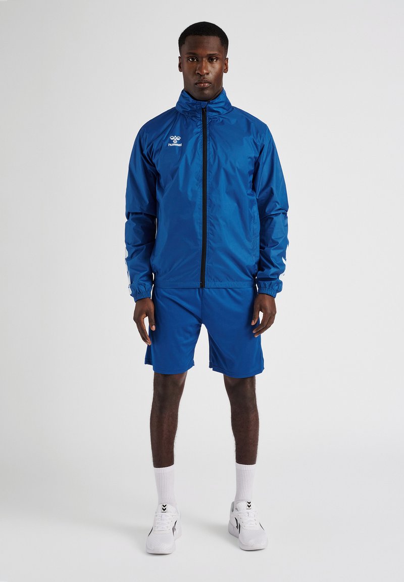 Mann steht und trägt eine blaue Sportjacke mit Reißverschluss, passende Shorts, weiße Socken und weiße Sportschuhe vor einem einfarbigen Hintergrund.