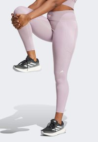 Leggings rosa chiaro con vita alta, design senza cuciture e tasche laterali, abbinati a scarpe da ginnastica nere e bianche con dettagli in rete.