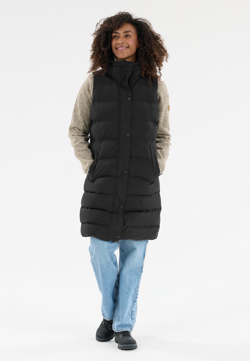 Svart purrig väst med hög krage och knappstängning, tillsammans med en grå långärmad topp, blå jeans och svarta stövlar.