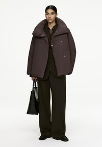 Puffer jacket in deep brown with a high collar and oversized fit; features large buttons. Paired with a black textured tote bag and loose trousers.