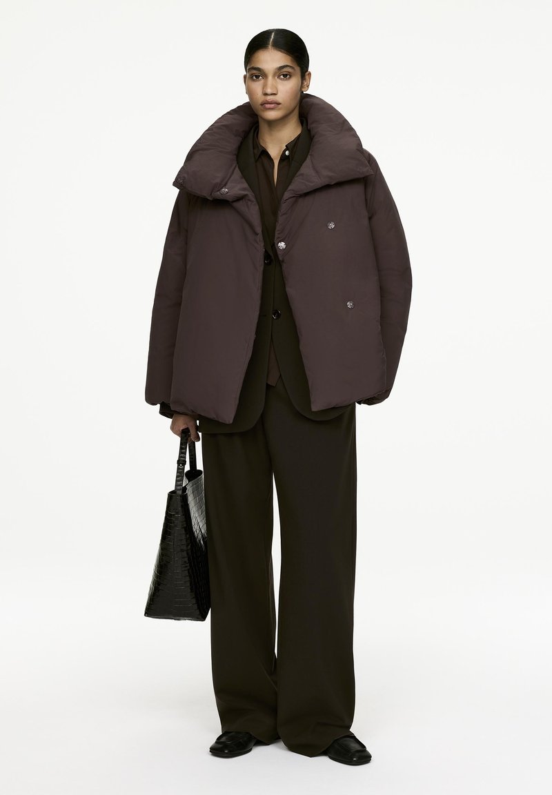 Puffer jacket in deep brown with a high collar and oversized fit; features large buttons. Paired with a black textured tote bag and loose trousers.