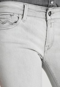 Jeans de mezclilla gris claro con un diseño clásico de cinco bolsillos, costuras visibles y un cierre estándar con botón y cremallera.