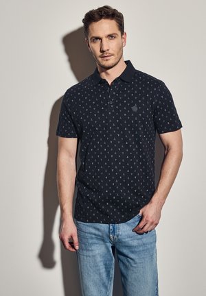Schwarzes Polohemd mit einem kleinen Segelbootmuster, kurzen Ärmeln und Kragen. Getragen mit hellblauen Jeans. Mit einem Logo auf der Brust.