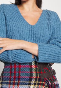 Sininen neulepusero, jossa on V-pääntie, tekstuuri kuvio ja puffihihat. Yhdistetty ruudulliseen hameeseen, jossa on punaisia, vihreitä ja keltaisia värejä.