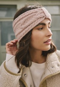 Bentiță roz tricotată cu model texturat în forma de cablu, purtată cu un pulover crem și o haină bej, având un finisaj curat și moale.