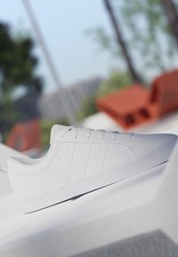 Zapatilla blanca de caña baja con cordones y sutiles rayas laterales colocada sobre una superficie blanca, fondo exterior desenfocado con árboles y sillas rojas.