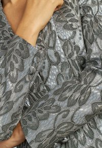 Tessuto metallico argento con motivi floreali intrecciati e testurizzati; presenta dettagli con bottoni sui polsini e una superficie liscia e sleek.