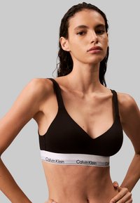 Bralette en coton noir avec un design en V, des bretelles larges et une bande blanche avec la marque "Calvin Klein" au bas.