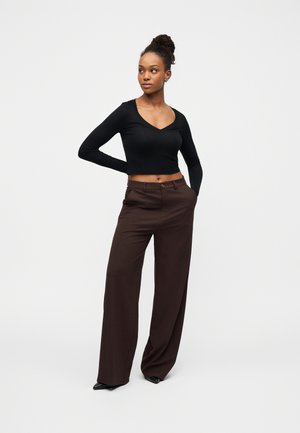 Kvinne med mørkt hår i knute, iført en svart langermet crop-top og mørkebrune vide bukser, står med hendene i lommene.