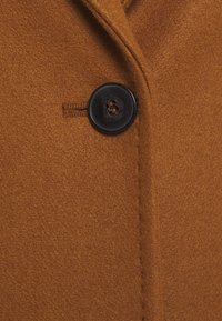 Tessuto di cappotto in lana marrone con una texture morbida, caratterizzato da un bottone in plastica nero e dettagli di cuciture visibili lungo la patta dei bottoni.