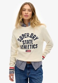 Pull en tricot crème avec le texte "SUPER DRY STATE ATHLETICS", présente des accents marine, des poignets rayés rouges, superposé à une chemise rayée bleue.