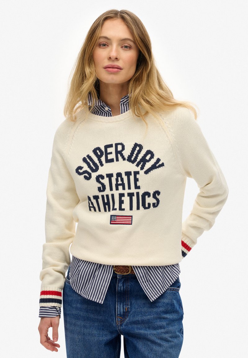 Pull en tricot crème avec le texte "SUPER DRY STATE ATHLETICS", présente des accents marine, des poignets rayés rouges, superposé à une chemise rayée bleue.