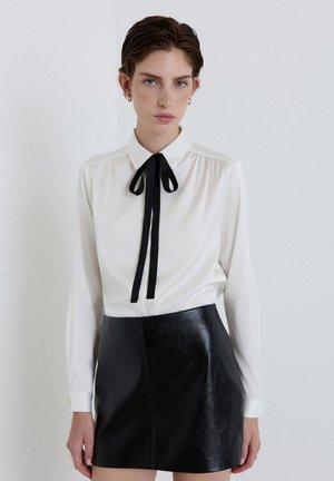 Donna che indossa una camicetta bianca con un papillon nero e una gonna di pelle nera, in piedi davanti a uno sfondo bianco uniforme.