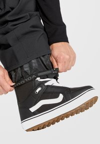 Bottes de neige noires avec des accents blancs, en tissu texturé et une semelle en caoutchouc, en train d'être attachées au-dessus de pantalons noirs avec un poignet zippé.