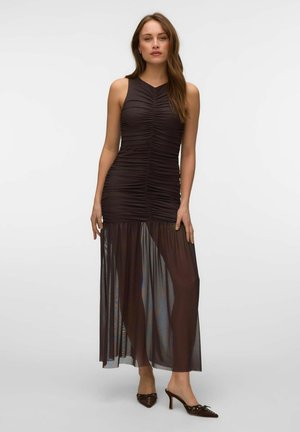 Femme portant une robe marron foncé sans manches froncée avec un panneau de jupe longue transparente et des chaussures à talons hauts noires pointues, debout.