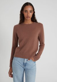 Frau mit langen, lockigen Haaren, die einen braunen Rundhalsausschnitt-Pullover und hellblaue, hochtaillierte Jeans trägt, posiert mit einer Hand in der Tasche.