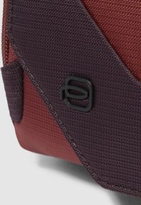 Bolsa de tela texturizada en burdeos y púrpura, con un logo negro, paneles contrastantes y cierre de cremallera.