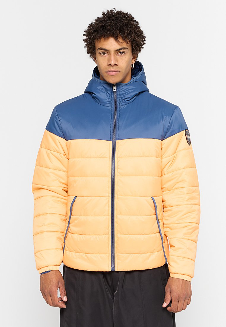 Uomo che indossa un giubbotto imbottito color block blu e pesca con cappuccio, in piedi contro uno sfondo grigio chiaro uniforme.