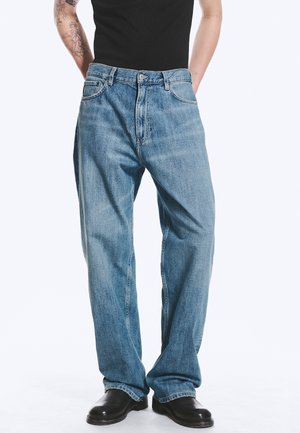 Personne portant un jean bleu clair ample, une chemise noire et des chaussures noires, debout avec les mains derrière le dos et un tatouage visible sur le bras gauche.